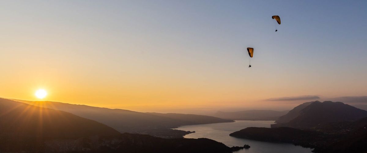 Parapentes en vol au-dessus du lac d'Annecy avec un coucher de soleil spectaculaire en arrière-plan, illuminant les montagnes et l'eau