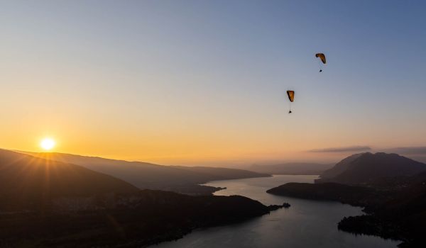 Parapentes en vol au-dessus du lac d'Annecy avec un coucher de soleil spectaculaire en arrière-plan, illuminant les montagnes et l'eau