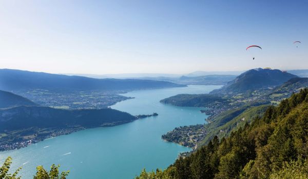 Vue du lac d'Annecy entouré de montagnes avec des parapentes en vol, un spot idéal pour les activités aériennes.