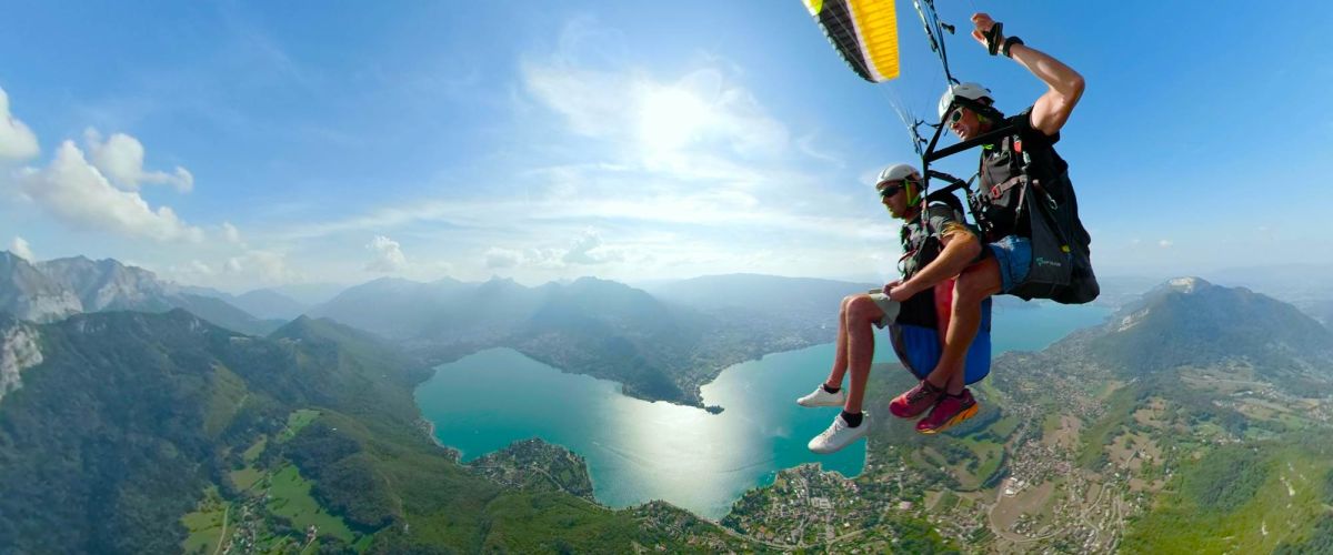 Deux personnes en parapente en tandem survolant le lac d'Annecy, avec un panorama spectaculaire des eaux et montagnes en arrière-plan