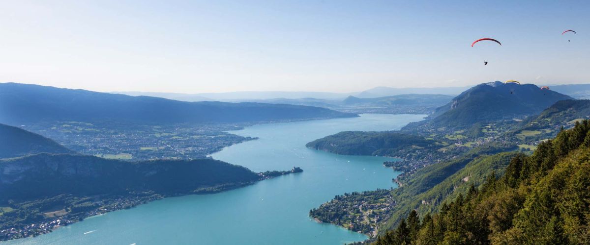 Vue du lac d'Annecy entouré de montagnes avec des parapentes en vol, un spot idéal pour les activités aériennes.